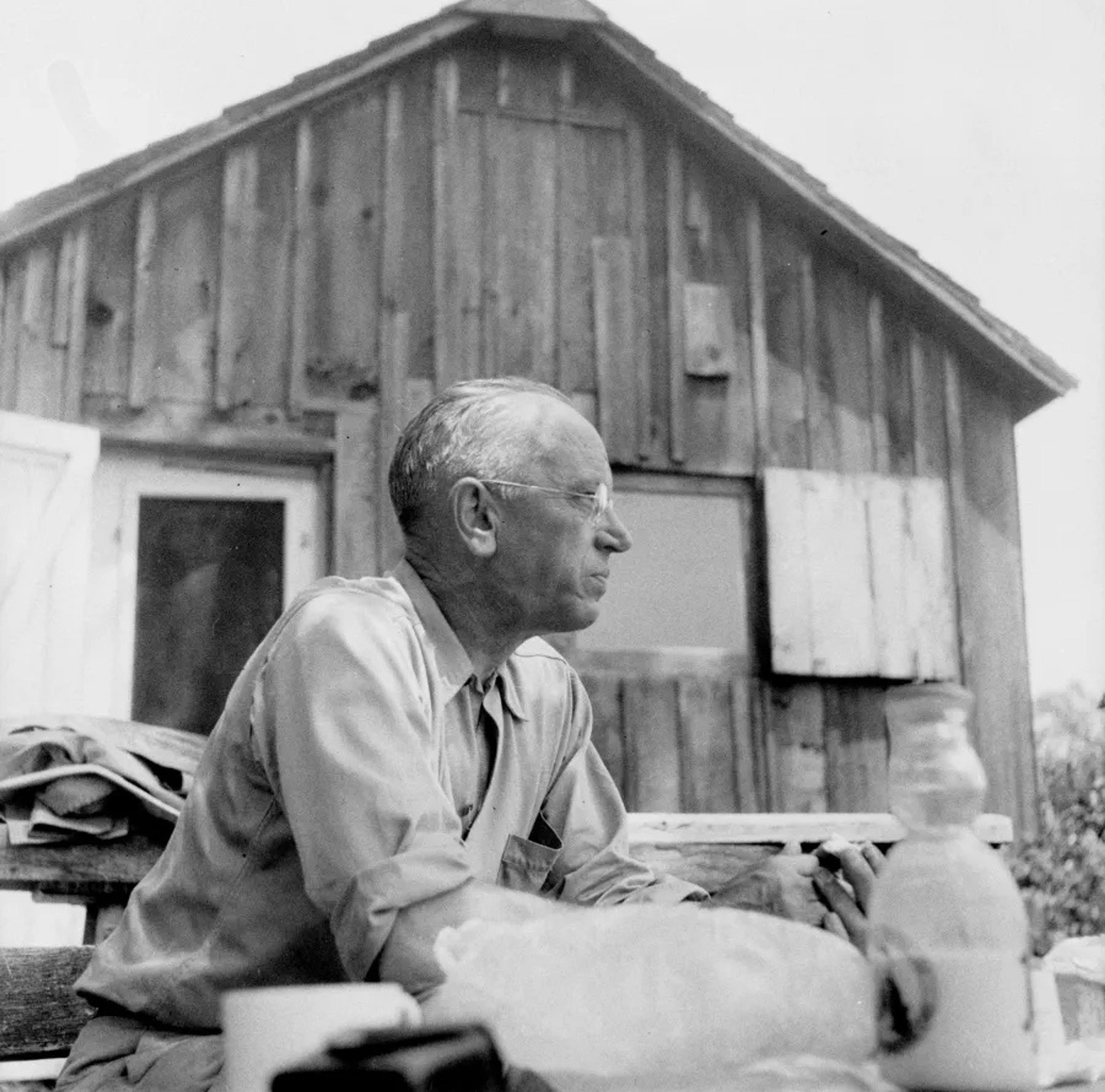 Black and white photo of an elderly man sitting outside a rustic wooden cabin, looking to the right with folded arms.