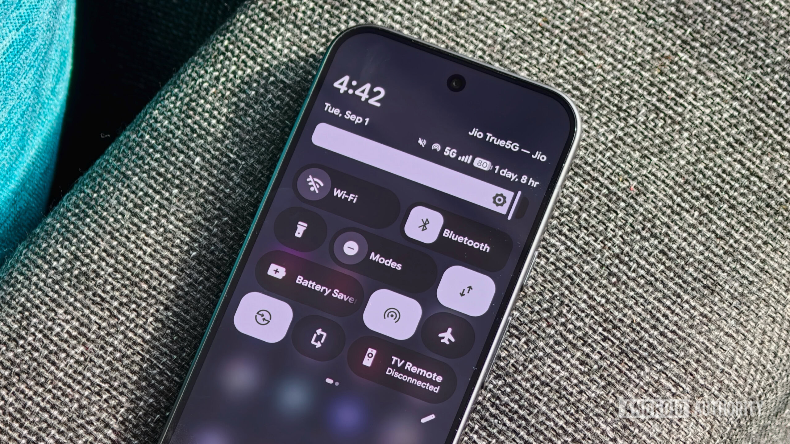 android quick settings tiles 1 An Android smartphone resting on a textured fabric surface displaying the expanded Quick Settings shade with tiles for Wi-Fi, Bluetooth, Modes, and Battery Saver