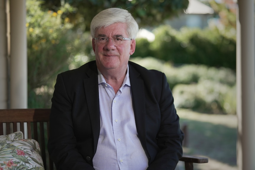 A man with grey hair and glasses sitting down.