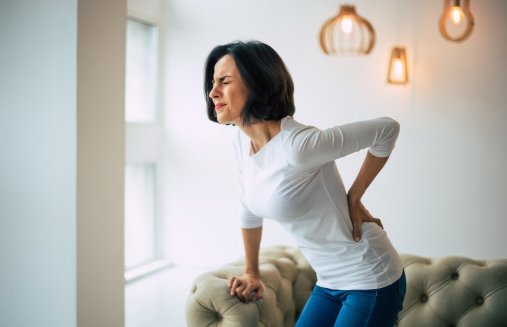 A woman holds her lower back in pain while leaning on a sofa.