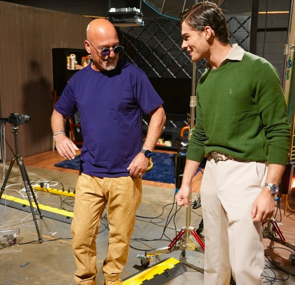 A bald man in sunglasses and a purple shirt talking to a man in a green sweater and beige pants.