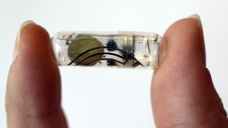 A bioresorbable battery in a capsule for transient ingestible bioelectronics, as held by two fingers on a white background.