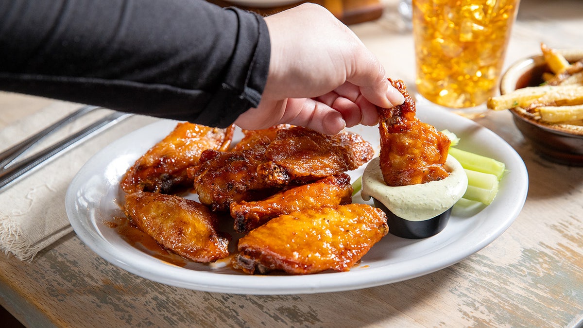 Hand holding a wing above plate of wings, dipping one into sauce.