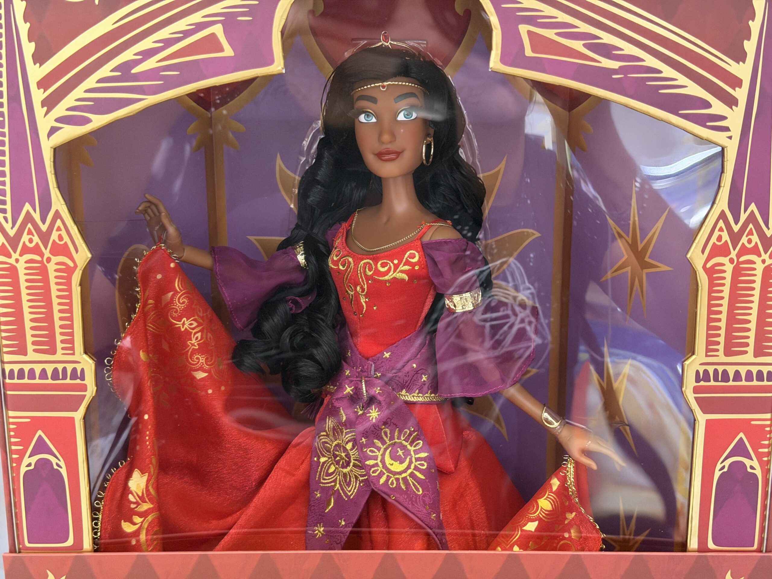 The bodice of her red dress is crafted of satin and embroidered with gold details. Her purple satin scarf is tied at her waist, proudly displaying gold star, flower, and moon designs. Sheer purple sleeves are banded with gold cuffs.
Her red skirt features a gold foil filigree pattern and gold thread detailing at the hem.
Her red skirt features gold foil filigree details and a gold detail at the hem.
