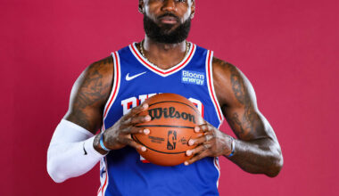 LeBron James, wearing a cap and casual jacket, speaks at an event, holding a microphone with a relaxed expression.