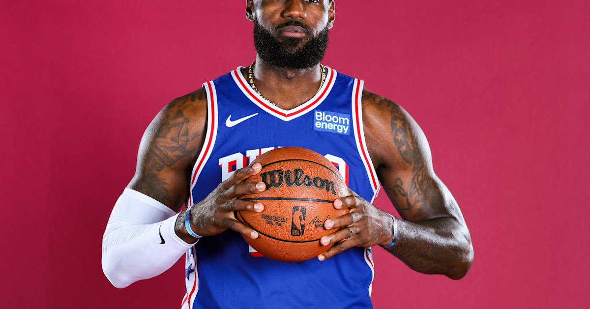 LeBron James, wearing a cap and casual jacket, speaks at an event, holding a microphone with a relaxed expression.