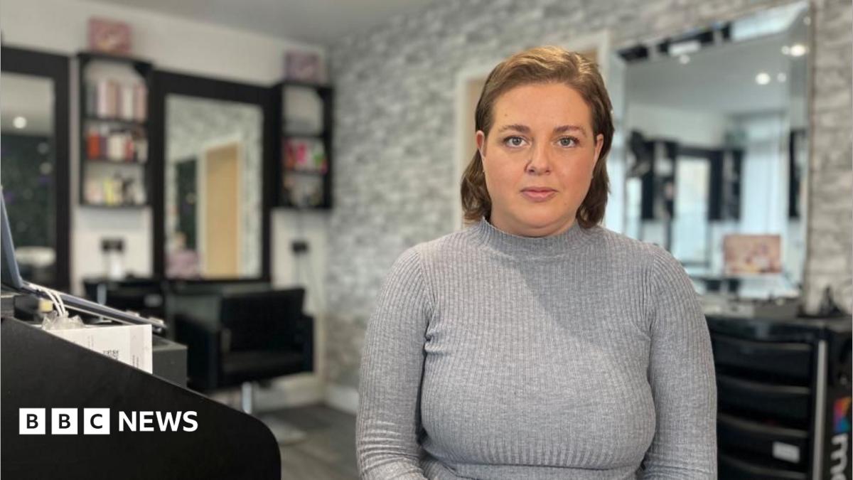 A brunette woman wearing a grey, ribbed turtleneck long sleeve top, facing the camera head on. She has light eyes and is stood in a beauty salon.