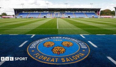 The Shrewsbury Town club crest with a stand at its home ground in the background