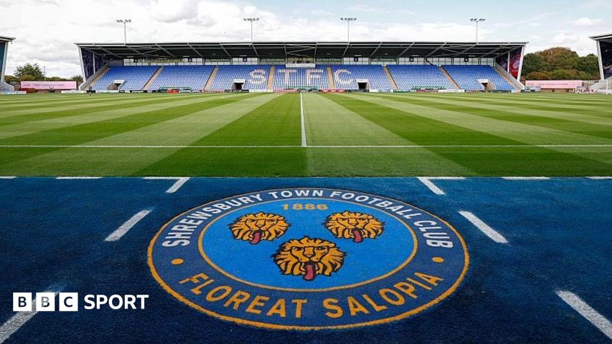 The Shrewsbury Town club crest with a stand at its home ground in the background