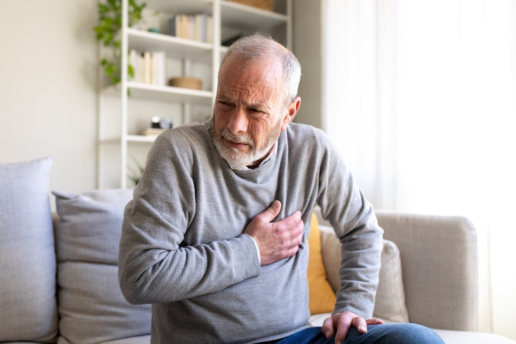 Elderly man clutching his chest in pain while sitting on a sofa.