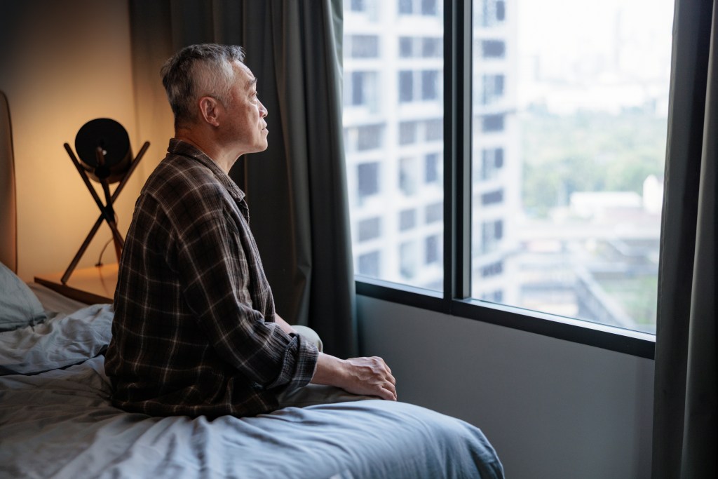 An elderly man in a plaid shirt sits on his bed, looking out a window at a distant city view.