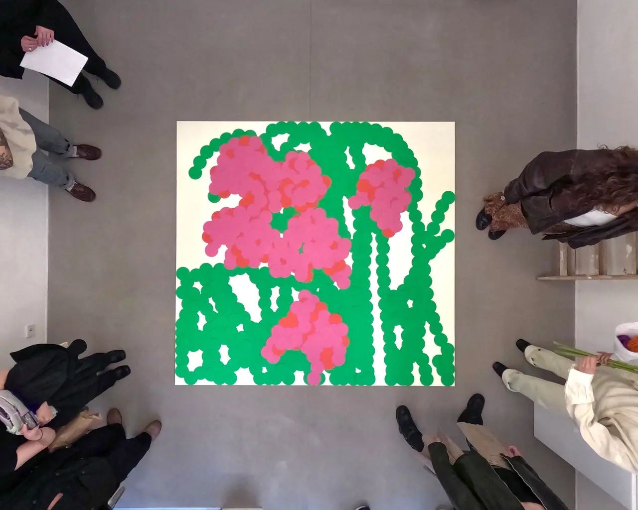 Vibrant hand-cut paper collage featuring bright pink flowers with green stems and leaves against a cream background, viewed from above with people standing around it