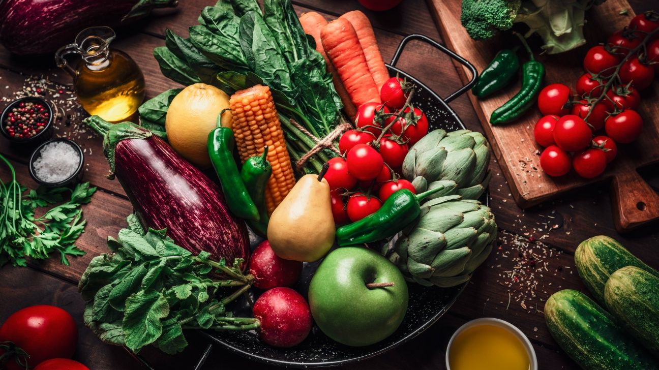 A bowl full of fruits and vegetables on a wooden table, surrounded by more fruits, vegetables, and spices
