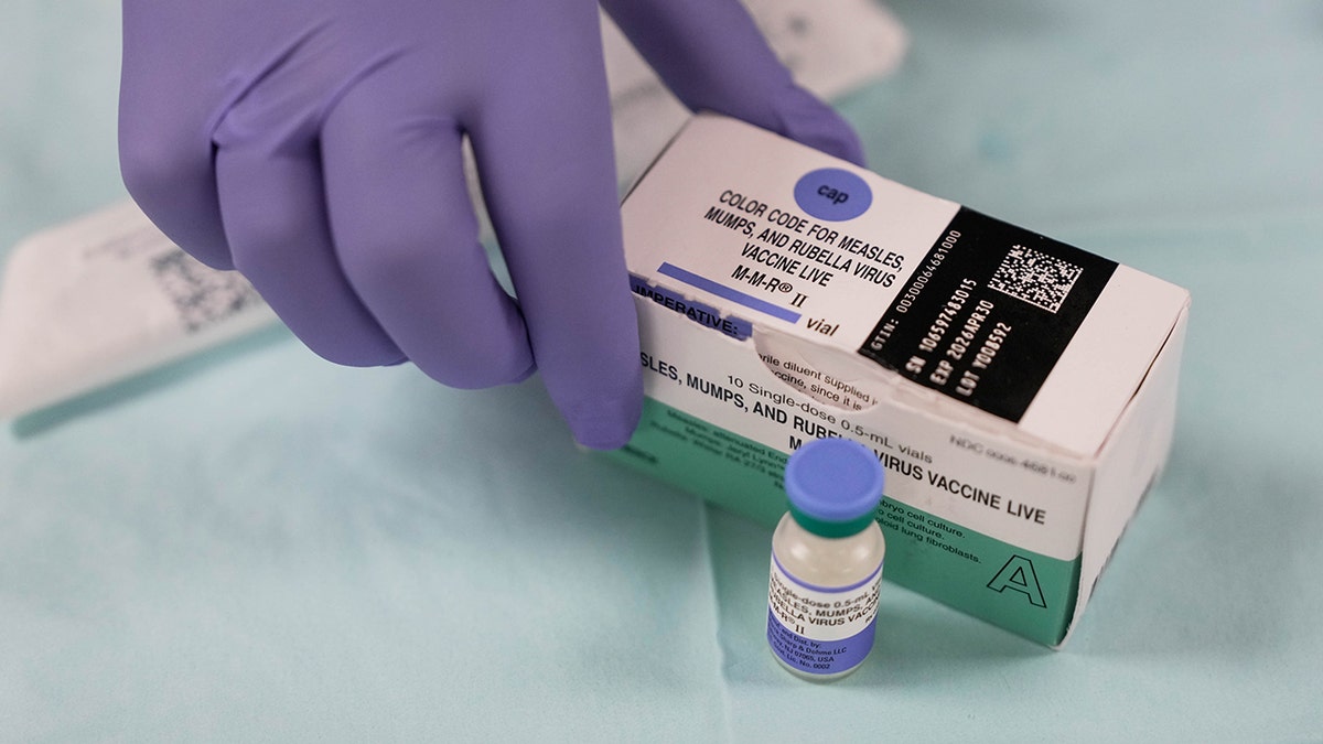 A box of the MMR vaccine, closeup of gloved hand holding box of the MMR vaccine next to a vial of the vaccine.