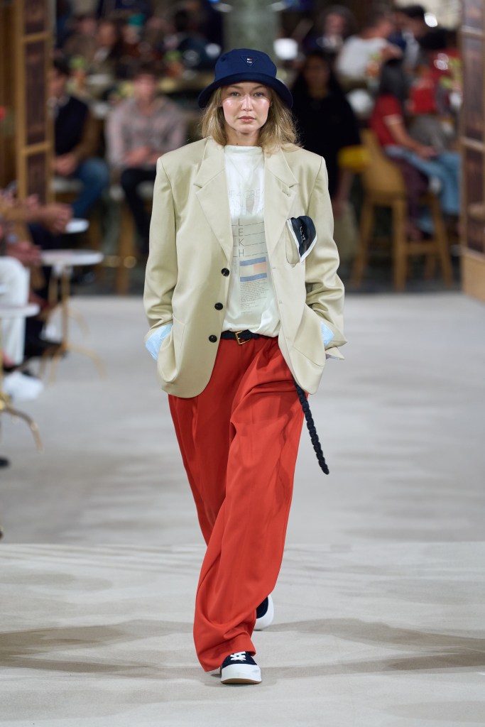 Gigi Hadid walks the runway in a beige blazer, white graphic t-shirt, orange wide-leg pants, and a navy bucket hat.