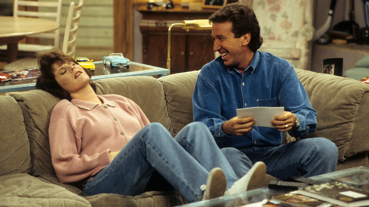 Patricia Richardson sits on the couch and throws her head back during an episode of "Home Improvment" as Tim Allen looks at her lovingly