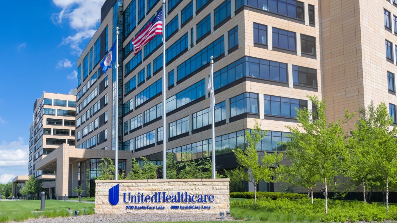 The UnitedHealth Group headquarters building features light-colored panels and blue-tinted windows across multiple stories, under a bright blue sky with scattered clouds. In the foreground, a stone monument sign reads 'UnitedHealthcare' with '9700 HealthCare Lane' and '9800 HealthCare Lane'. An American flag, a blue flag with a seal (likely Minnesota), and a white flag fly from three tall flagpoles. Green trees and manicured lawns surround the building's entrance.