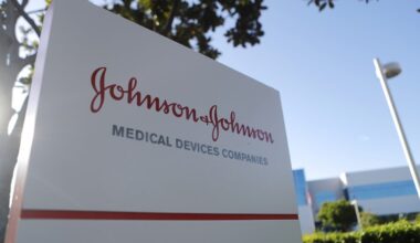 A close-up, low-angle shot of a white rectangular corporate sign for Johnson & Johnson. The company's name is written in a distinctive red script, with 'MEDICAL DEVICES COMPANIES' in a smaller, gray sans-serif font below. Sunlight creates a bright lens flare on the left side of the sign, with blurred green tree foliage and a clear blue sky in the background. A white light pole is visible on the right, and parts of a modern building with blue-tinted windows can be seen in the distance.