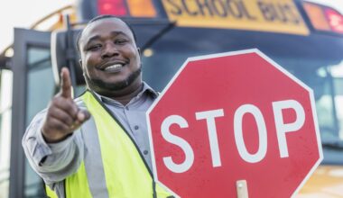 A smiling Black man, wearing a bright yellow reflective safety vest over a gray collared shirt, holds a red octagonal stop sign in his right hand. He points forward with his left index finger. Behind him, a yellow school bus is visible, with the top part of the 'SCHOOL BUS' text on its front blurred in the background. The man has a beard and looks directly at the camera with a friendly expression.