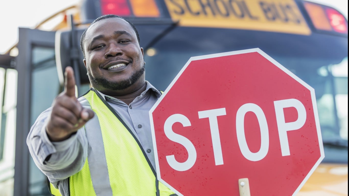 A smiling Black man, wearing a bright yellow reflective safety vest over a gray collared shirt, holds a red octagonal stop sign in his right hand. He points forward with his left index finger. Behind him, a yellow school bus is visible, with the top part of the 'SCHOOL BUS' text on its front blurred in the background. The man has a beard and looks directly at the camera with a friendly expression.