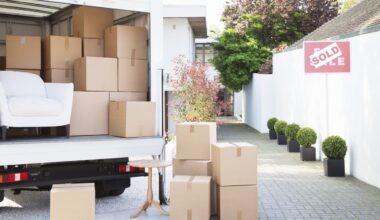 The back of an open white moving truck is seen, filled with numerous brown cardboard boxes and a white upholstered armchair. Outside the truck, a small wooden side table and several stacks of packed boxes are on a paved driveway, leading to a modern white house. A red 'SOLD' sign is visible to the right, in front of a white wall lined with potted green shrubs.