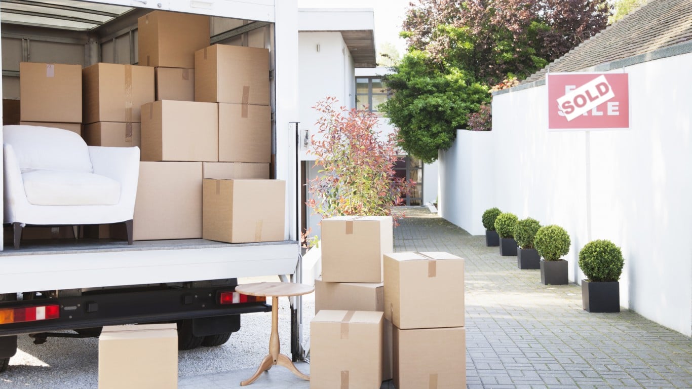 The back of an open white moving truck is seen, filled with numerous brown cardboard boxes and a white upholstered armchair. Outside the truck, a small wooden side table and several stacks of packed boxes are on a paved driveway, leading to a modern white house. A red 'SOLD' sign is visible to the right, in front of a white wall lined with potted green shrubs.