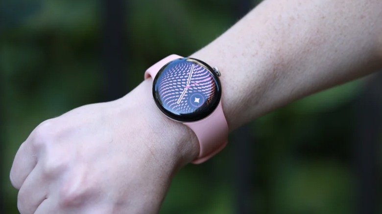 Photo of a Google Pixel 5 smartwatch with a pink band and an abstract pink and black face worn on a wrist with hand in a fist..