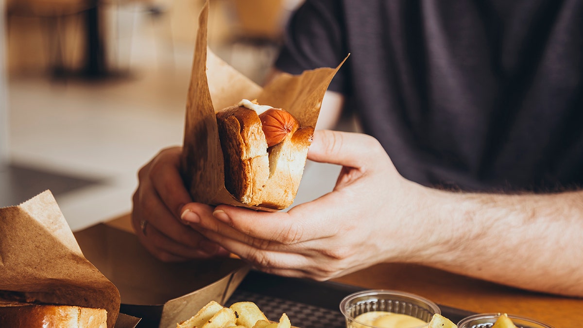 Man's hands seen up close as he holds hot dog in bun and paper.