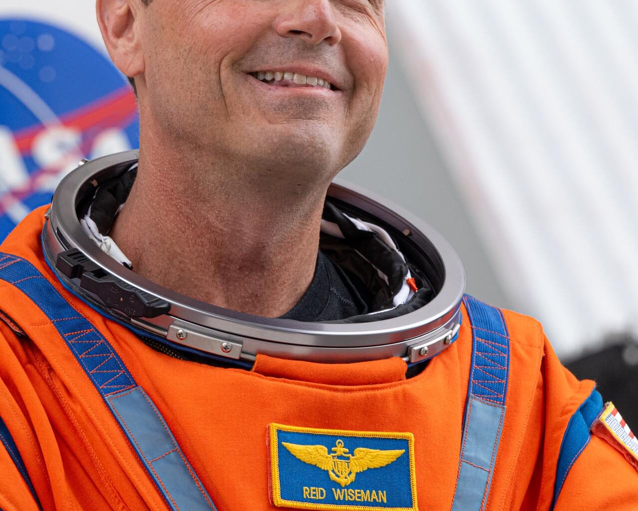 Reid Wiseman looks toward supporters, smiling, wearing an orange spacesuit with his name and a NASA meatball patch.