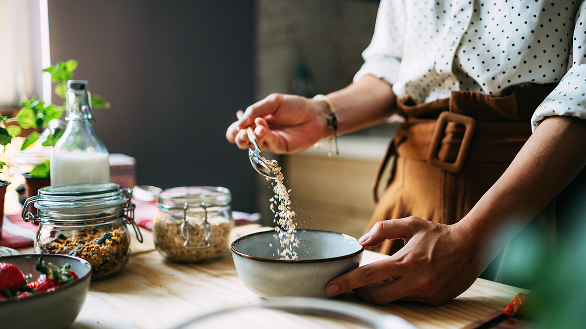 woman in kitchen pours dried oats into a bowl from a spoon