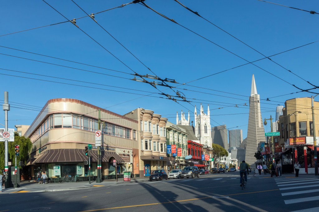 Old houses and businesses line a San Francisco street with the Transamerica Pyramid in the background.