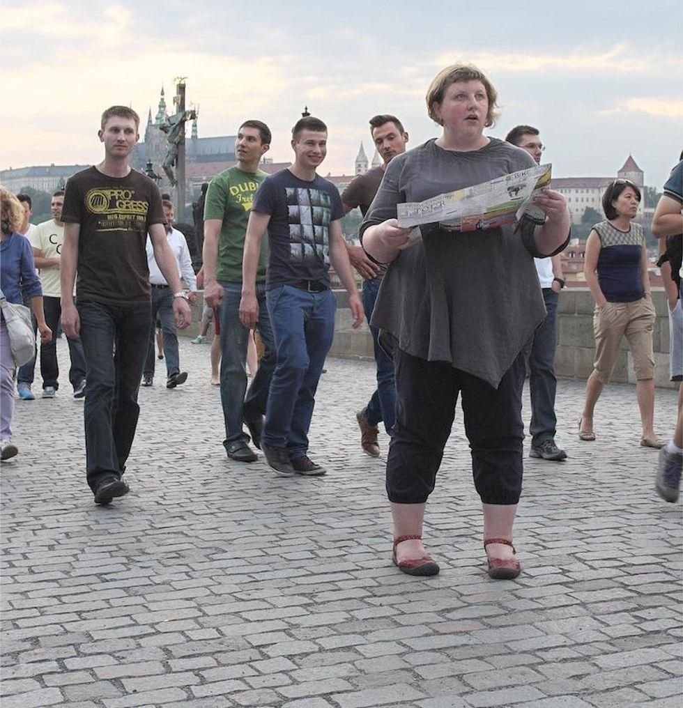Four young men walking behind a heavy set woman on a cobblestone street, laughing at a heavy set woman behind her back.
