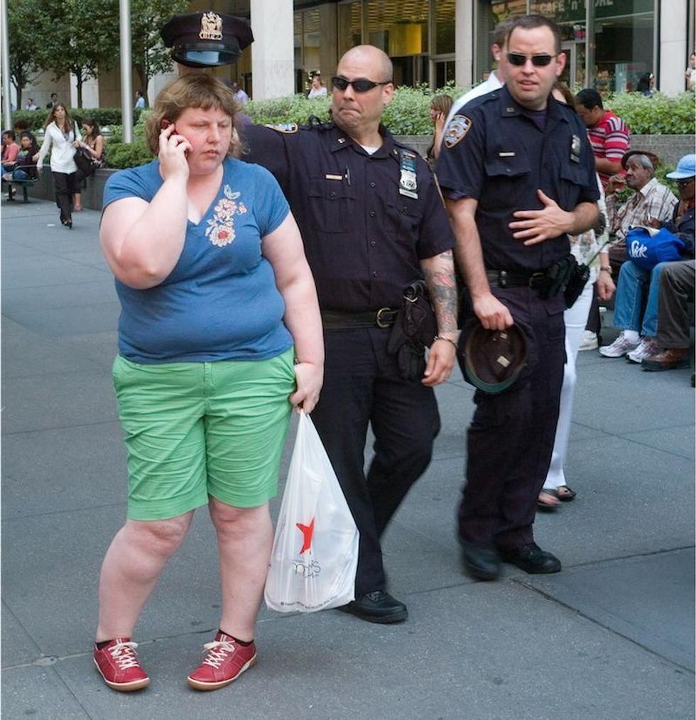 A policeman in uniform standing behind a heavy set woman who is on her phone, and holding his police hat above her head in mocking fashion.