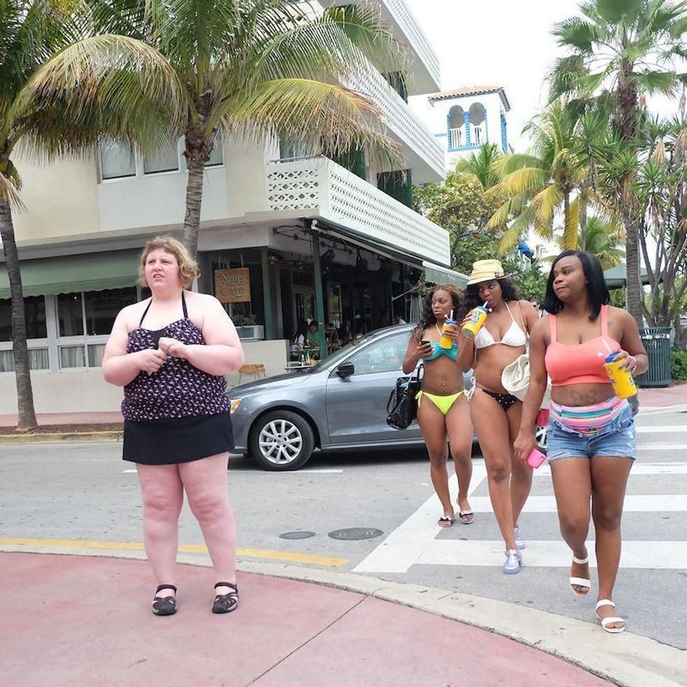 Three women in bikinis crossing a street, looking at a heavy set woman.