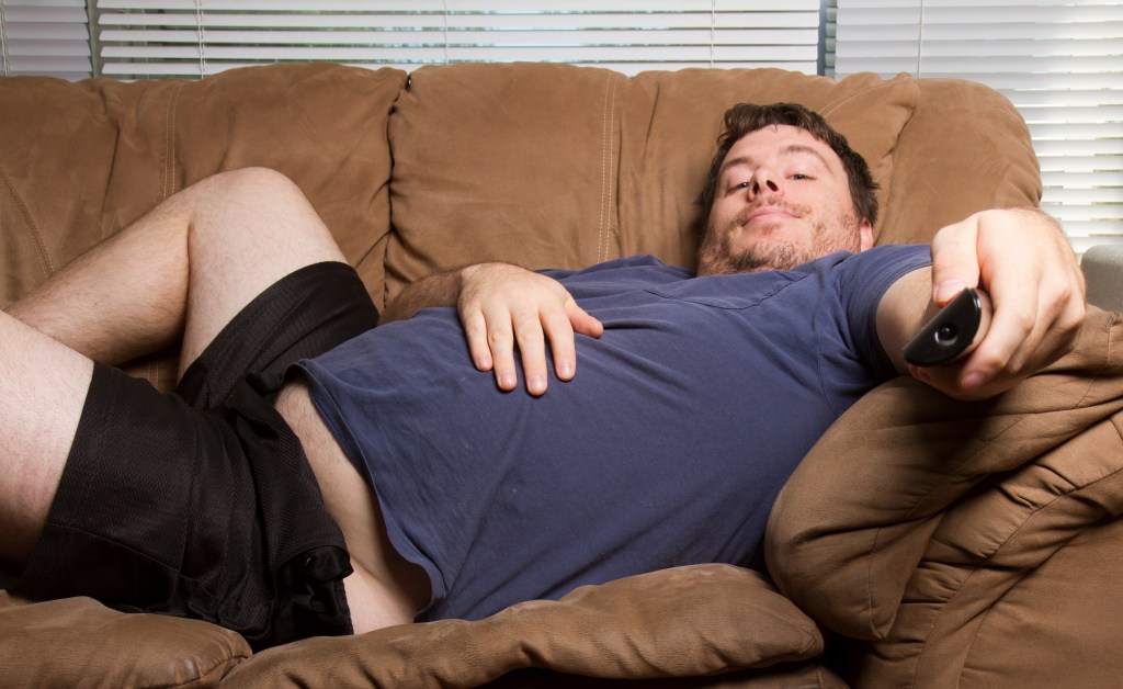 An overweight man lying on a couch holding a TV remote.
