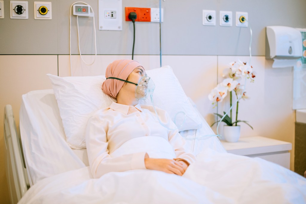 An Asian female cancer patient wearing an oxygen mask and a headscarf, lying in a hospital bed.