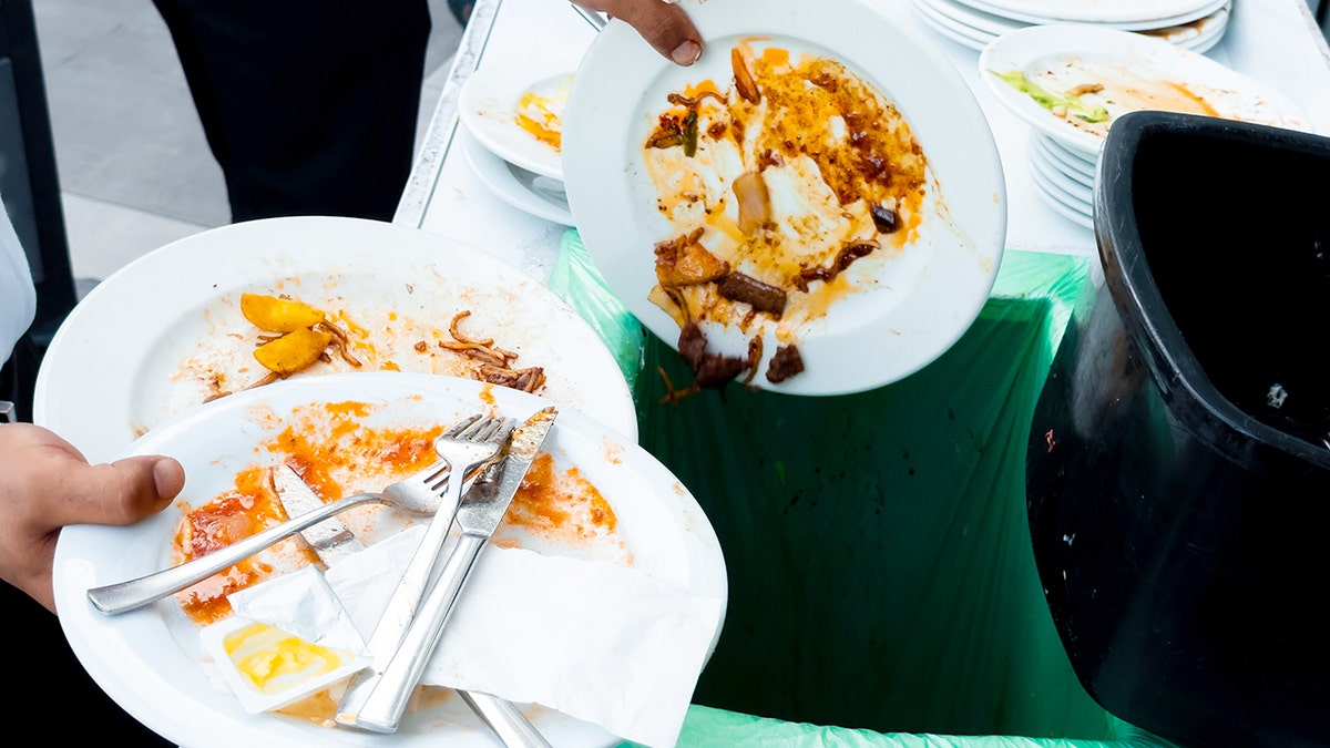 A restaurant server dumps food from dirty plates into the garbage.