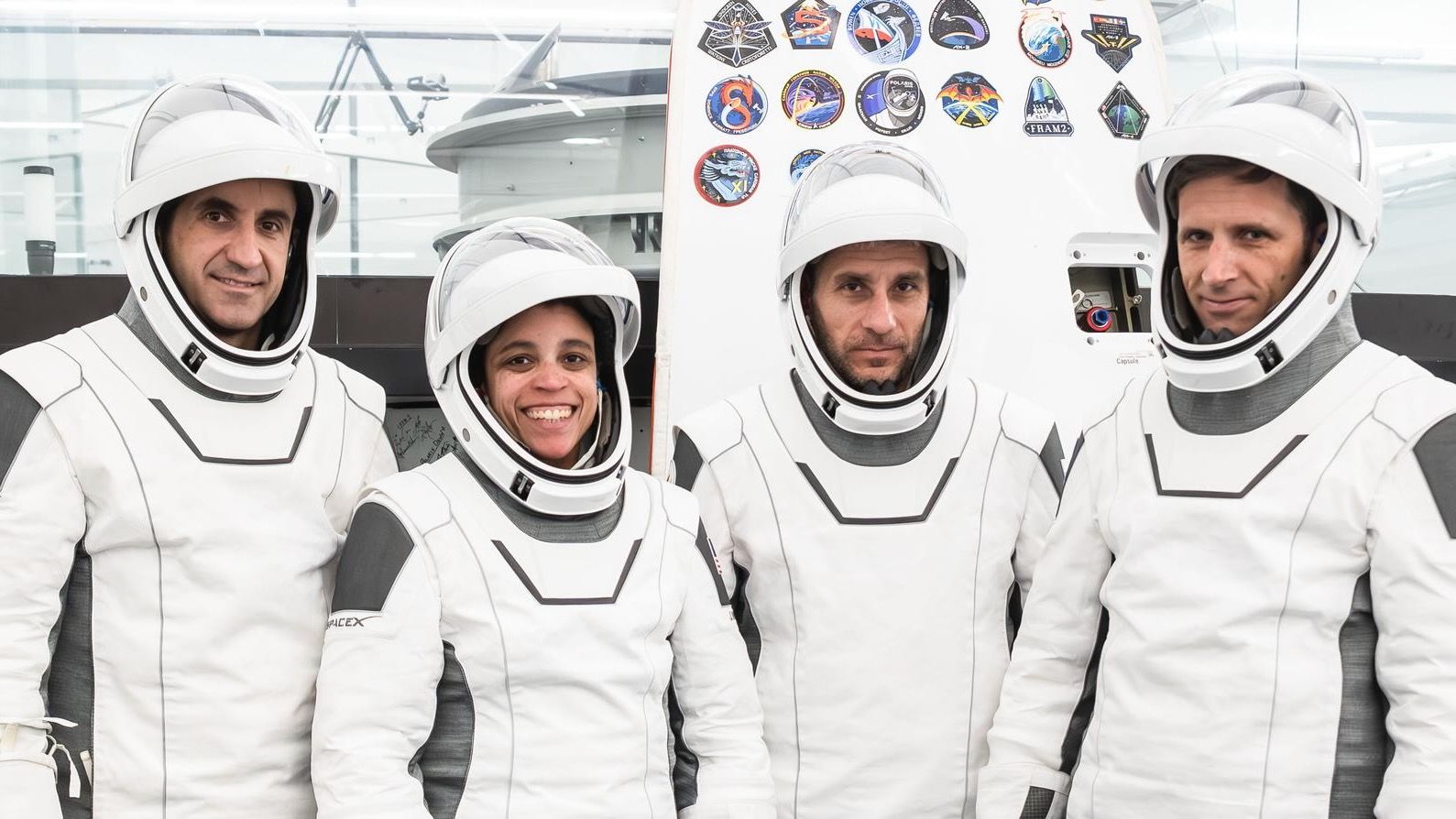 three male and one female astronauts in white flight suits stand in a big, white-walled room