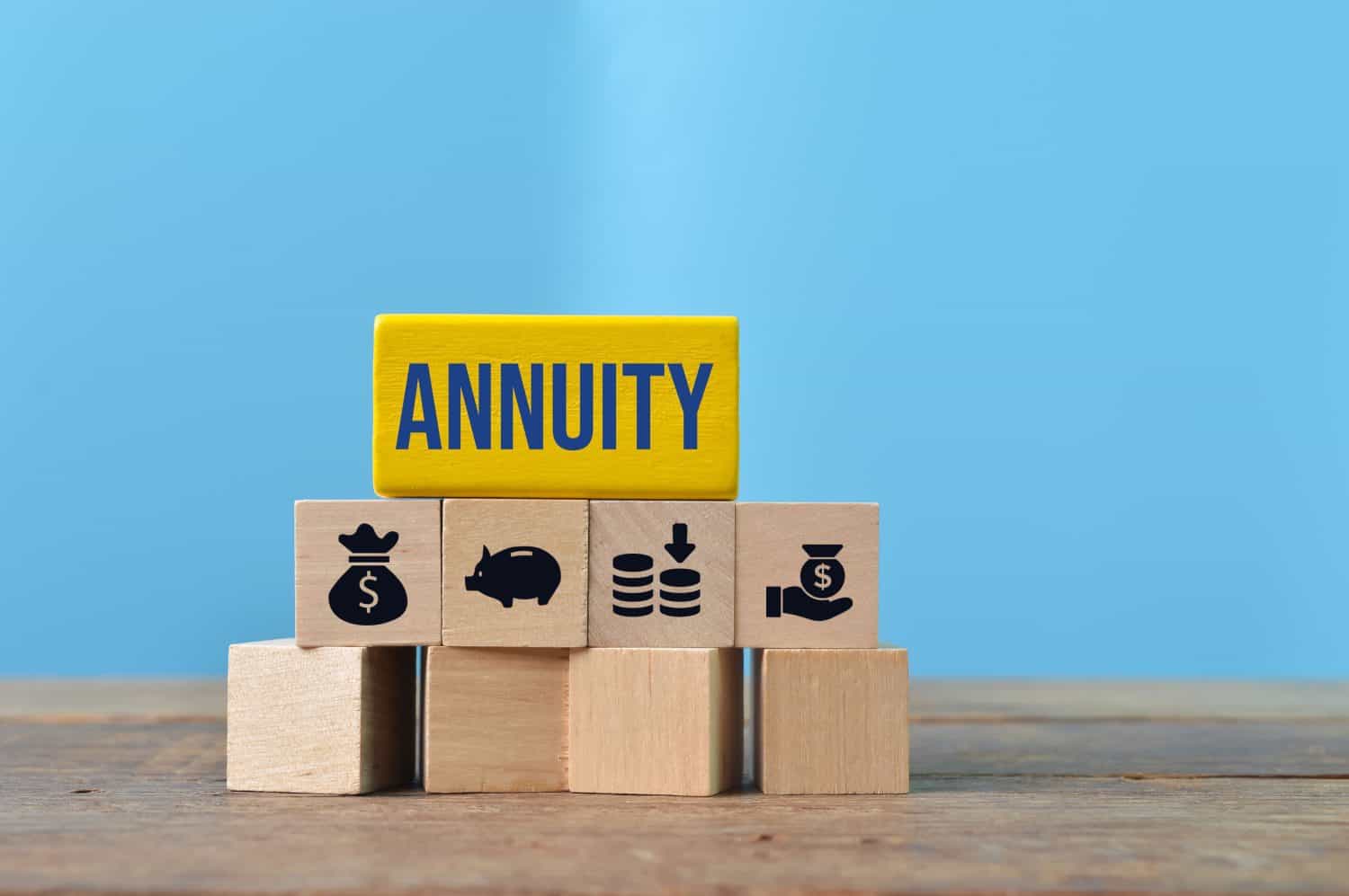 A yellow wooden block with the word 'ANNUITY' in blue text is stacked on four smaller natural wood blocks. These bottom blocks feature black icons: a money bag with a dollar sign, a piggy bank, stacks of coins with a downward arrow, and a hand holding a dollar bill. Below these are three empty natural wood blocks, all set on a wooden surface against a light blue background.