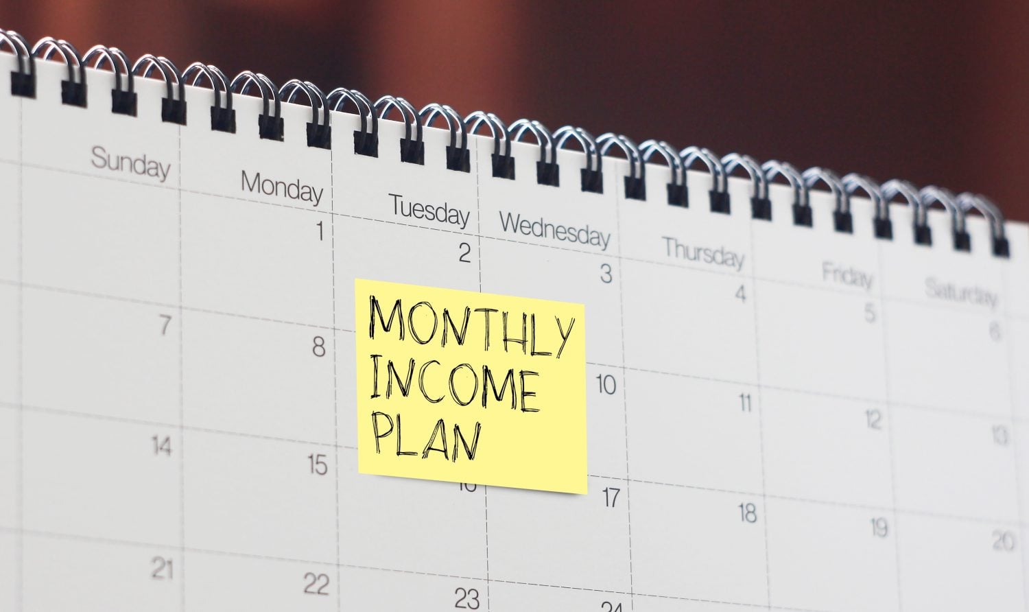 A close-up shot of a white desk calendar with visible dates and days of the week. A bright yellow square sticky note is prominently placed on the date '8', displaying the handwritten text 'MONTHLY INCOME PLAN' in black capital letters. The metal spiral binding of the calendar is visible along the top edge.