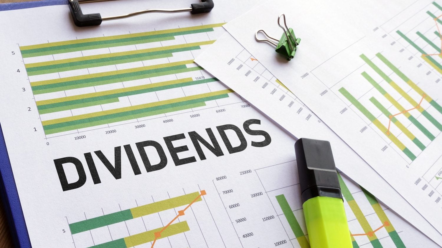 A close-up shot of financial documents on a blue clipboard, with the word 'DIVIDENDS' in large black letters across the center. Several green and yellow bar graphs, some with orange line overlays, are visible on the papers. A green binder clip is on the top right paper, and a bright yellow-green highlighter rests on the bottom right.