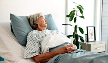 An elderly woman with short grey hair and a pensive expression lies in a hospital bed. She is wearing a patterned patient gown, propped up by a light blue pillow, with her hands resting on a light blue blanket. A green plant and a framed photo are on a bedside table in the soft background.