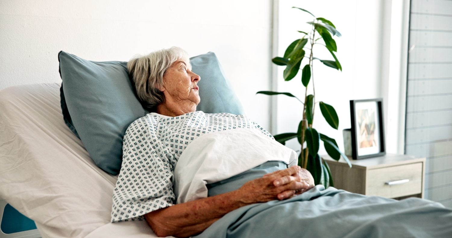 An elderly woman with short grey hair and a pensive expression lies in a hospital bed. She is wearing a patterned patient gown, propped up by a light blue pillow, with her hands resting on a light blue blanket. A green plant and a framed photo are on a bedside table in the soft background.