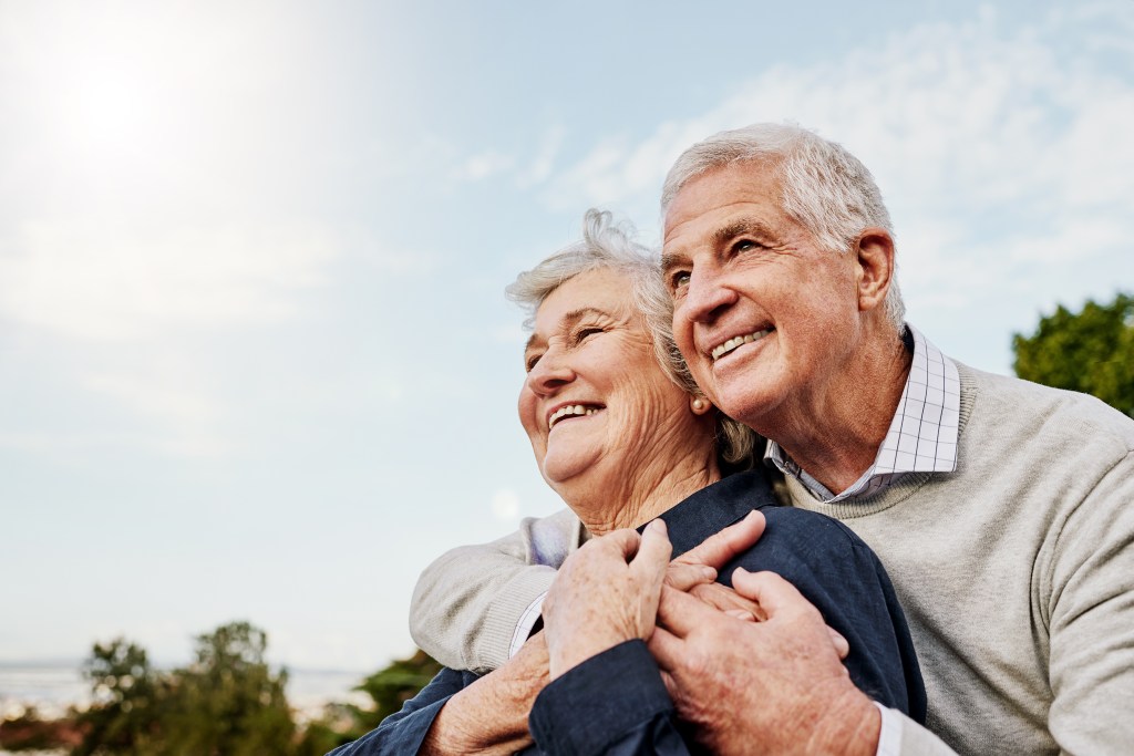An elderly couple embraces outdoors, smiling and looking into the distance.