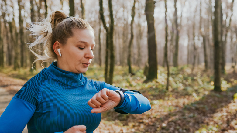 A woman jogging in the woods, looking at a watch on her wrist with wireless earphones attached