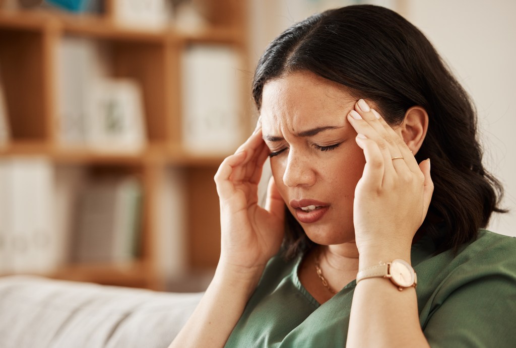 A woman sits on a sofa, grimacing in pain as she holds her hands to her temples, suggesting a headache or migraine.