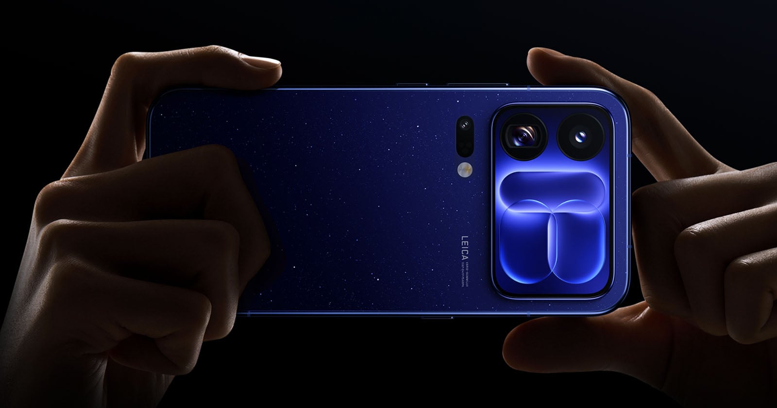 A pair of hands holds a dark blue smartphone with a starry pattern on the back and a prominent camera module featuring two large lenses and a distinct, glowing blue geometric design.