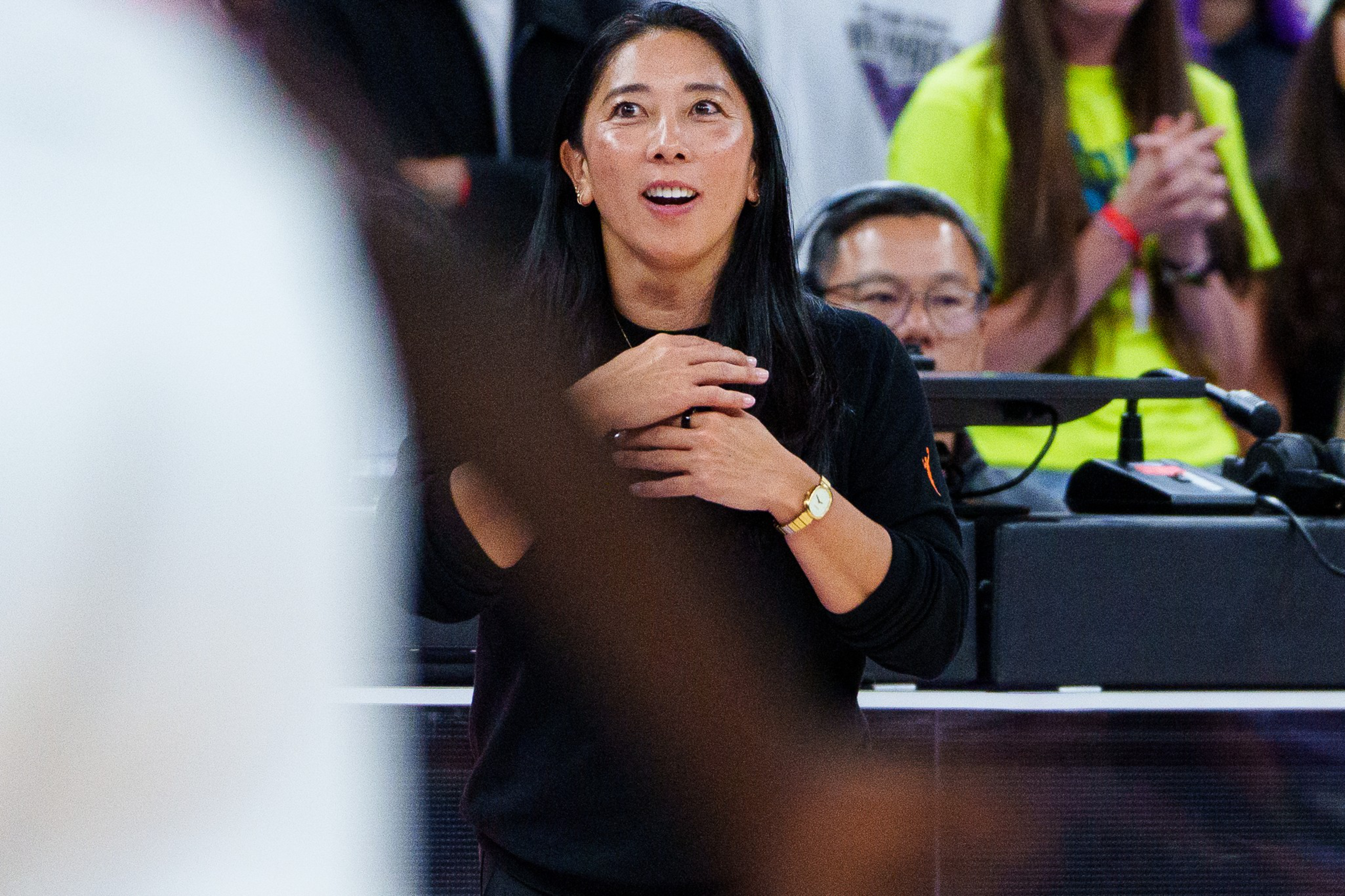 A woman with long black hair and a black sweater clasps her chest, looking upwards with a surprised or emotional expression.