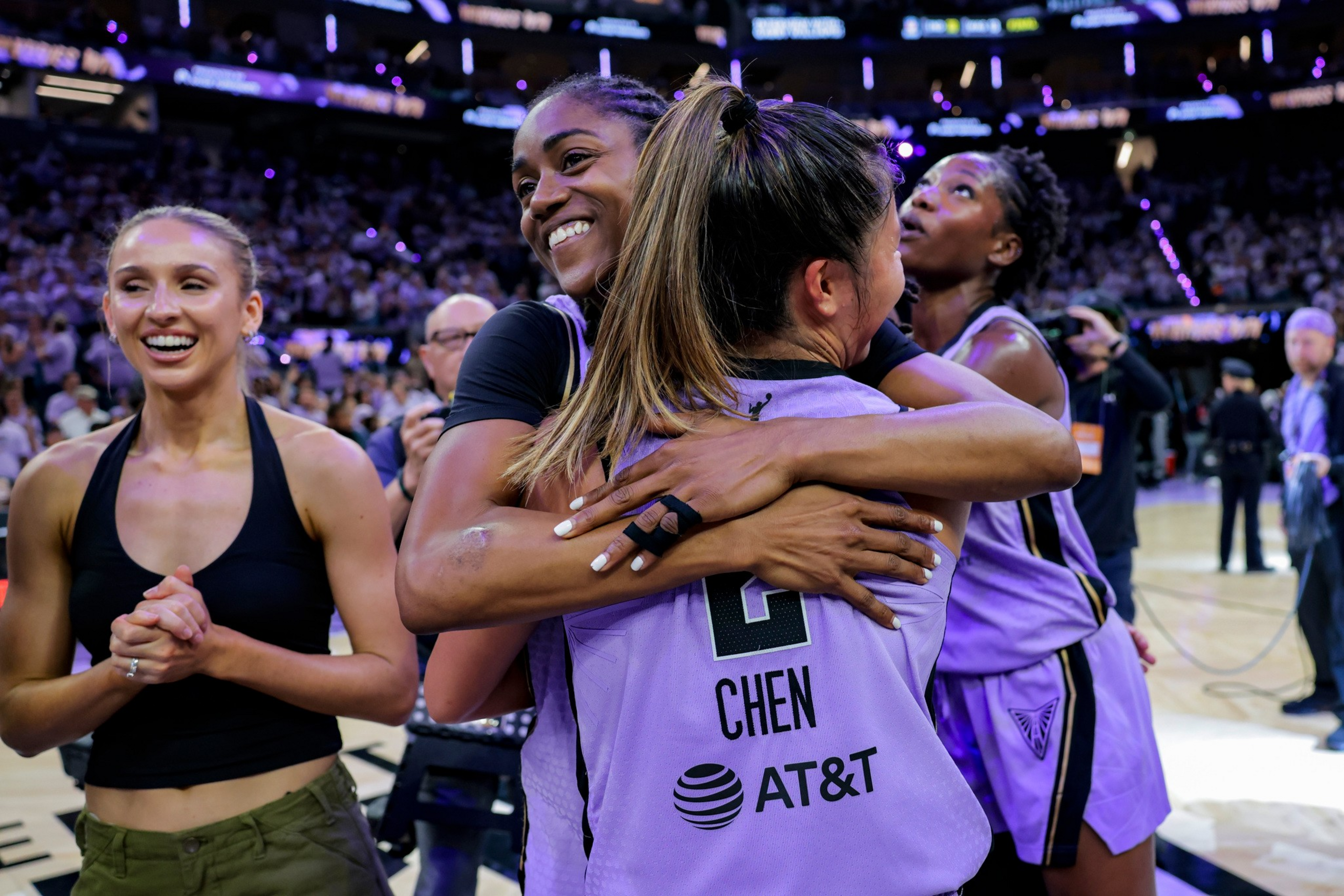 Two basketball players in purple jerseys hug while smiling, with one wearing the name “Chen,” as a woman in casual clothes smiles nearby on the court.