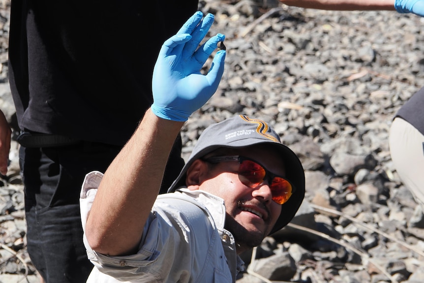 A man in a bowl hat and sunglasses smiles as he holds up a small snail shell